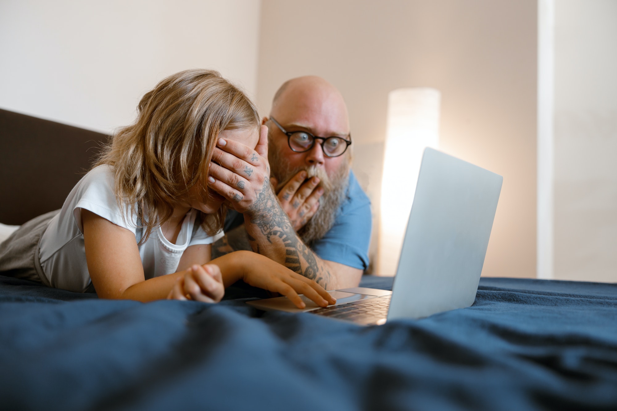 Shocked father closes eyes of daughter to protect from dangerous content on laptop on bed